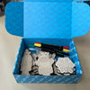 Open blue box with decorative patterns containing gray clay models and colorful tools on a neutral background.